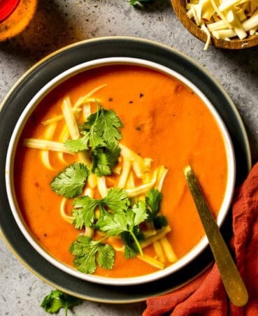 Smooth carrot soup in a shallow white bowl set on a plate. Shredded cheese and cilantro sprinkled over soup.