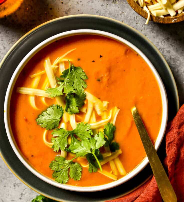 Smooth carrot soup in a shallow white bowl set on a plate. Shredded cheese and cilantro sprinkled over soup.