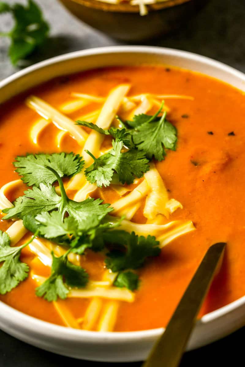 Smooth carrot soup in a shallow white bowl set on a plate. Shredded cheese and cilantro sprinkled over soup.