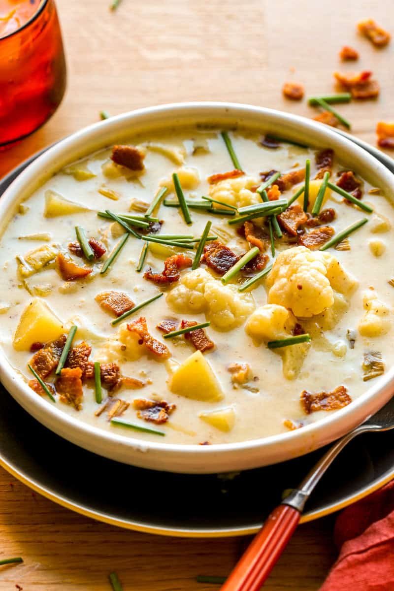 Creamy soup with chunks of cauliflower, potatoes, and leeks, in a shallow white bowl topped with bacon bits and chives.