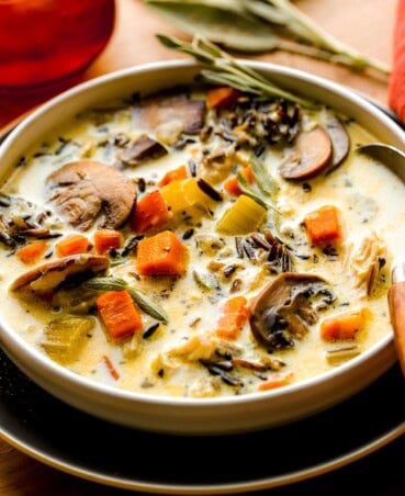 Chicken wild rice mushroom soup in a shallow white bowl set on a blue plate with a wooden-handled spoon set in it. Orange glass and fresh sage set on the counter next to it.