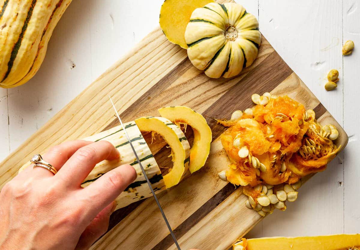 A set of hands slicing delicata squash on a wood cutting board into thin half moon slices.