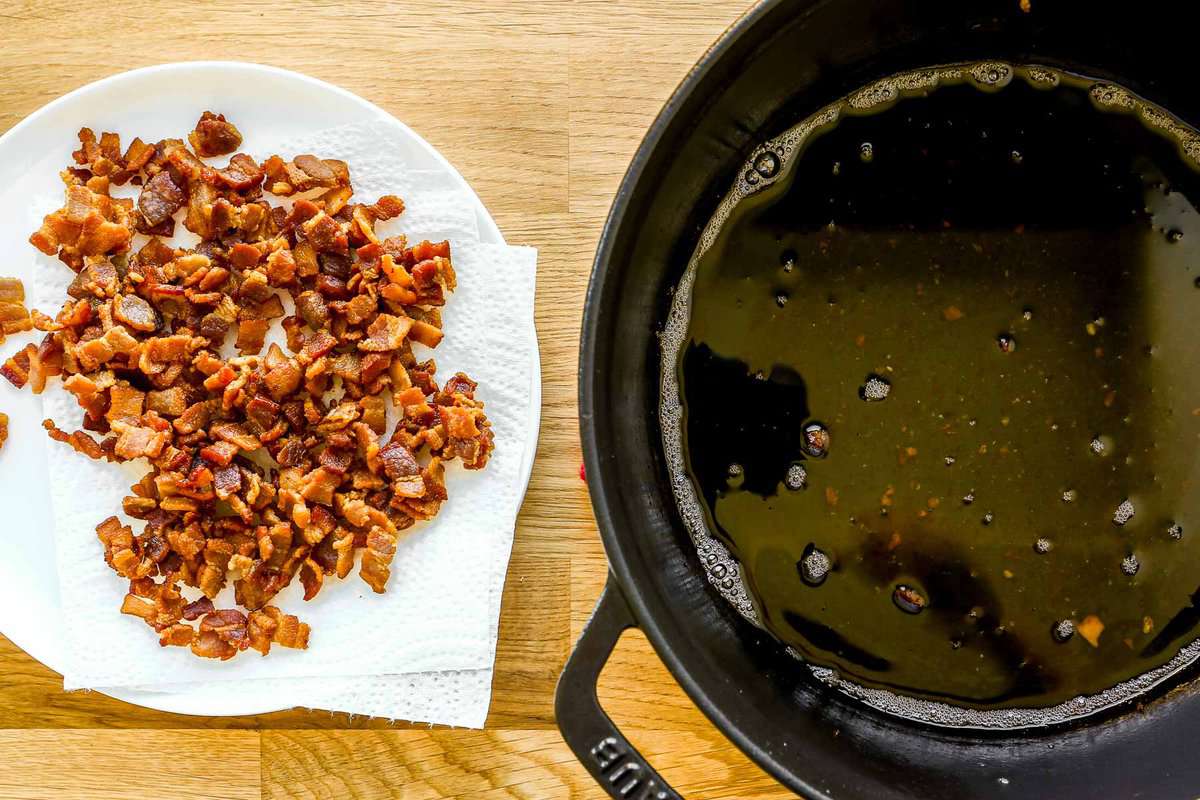 A paper-towel-lined plate with crispy bacon bits on it with a pot of bacon drippings set near by.