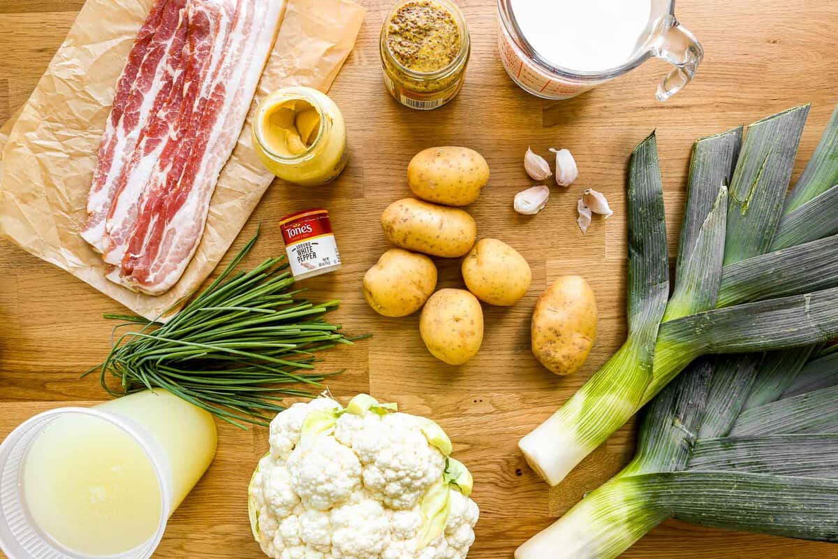 Slices of bacon, broth, chives, Dijon mustard, stone ground mustard, gold potatoes, leeks, a head of cauliflower, milk and 4 garlic cloves set out on a counter. 