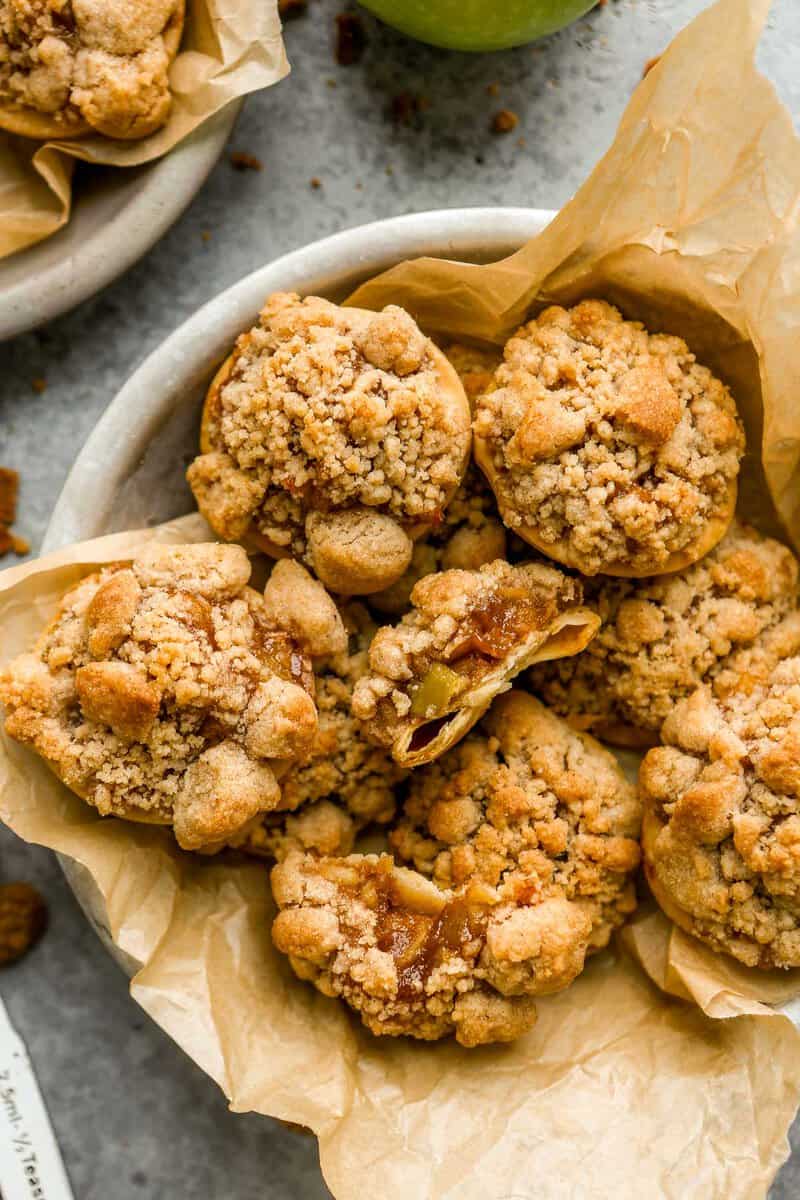 Apple pie cookies (pie crust rounds topped with apple pie filling and topped with streusel) arranged in a parchment-lined pie plate. One cookie set in the middle with a bite taken out of it.