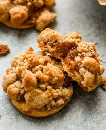 Three apple pie cookies set on a counter, two stacked on each other, the top cookie with a bite taken out showing the flaky pie crust, apple pie filling and streusel.