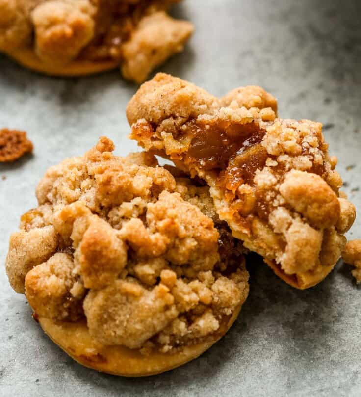 Three apple pie cookies set on a counter, two stacked on each other, the top cookie with a bite taken out showing the flaky pie crust, apple pie filling and streusel.