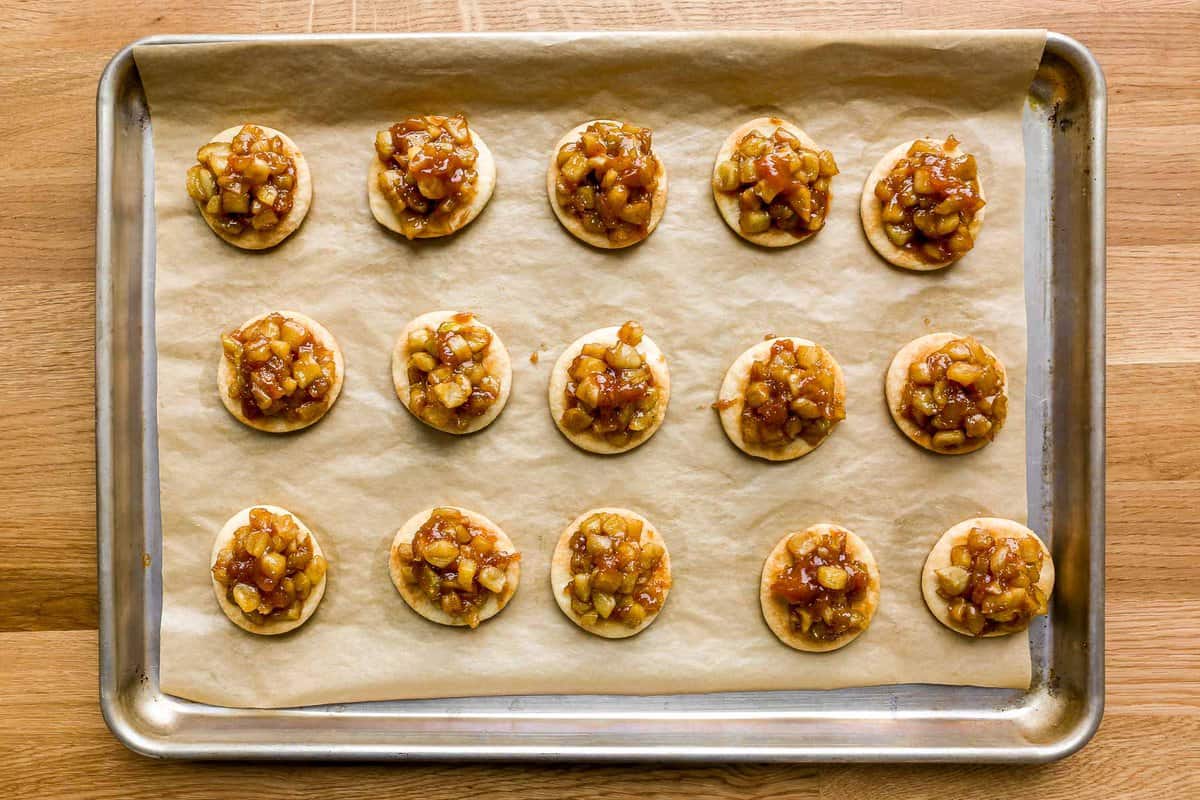 Baked pie crust rounds topped with an apple pie filling, set on a parchment-lined baking sheet.