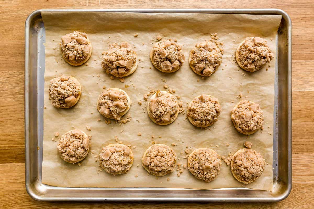 Baked pie crust rounds topped with an apple pie filling and a chunky streuse mixture, set on a parchment-lined baking sheet.