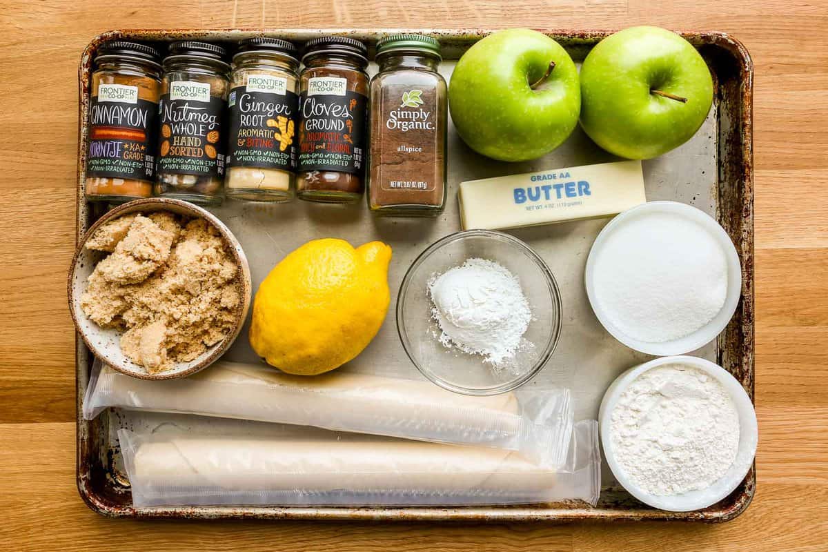 Bottled spices, Granny smith apples, brown sugar, lemon, cornstarch, sugar, pre-made pie crusts, and flour all measured out and set on a sheet tray.