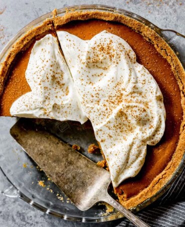 Pumpkin pie with graham cracker crust set on a blue counter with whipped cream spread over pie and dusted with cinnamon. A few slices taken out of pie and a serving spatula, nutmeg, grater, blue dessert plates and forks set off to the side.