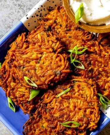 Crispy sweet potato fritters stacked on a blue and white speckled tray with a wooden bowl filled with yogurt off to the side. Sliced scallions scattered over top.