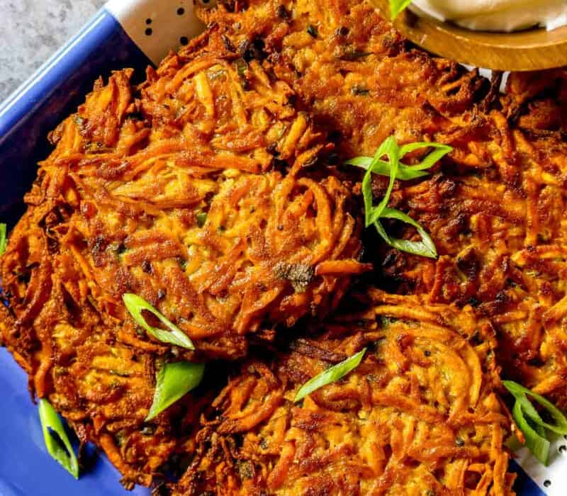 Crispy sweet potato fritters stacked on a blue and white speckled tray with a wooden bowl filled with yogurt off to the side. Sliced scallions scattered over top.