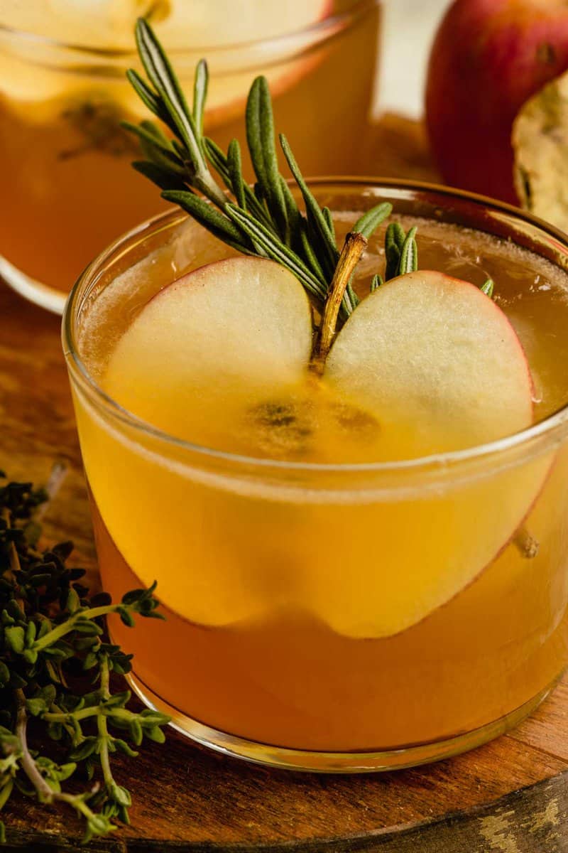 Short cocktail glass filled with an apple cider cocktail and garnished with an apple slice ad rosemary sprig. 