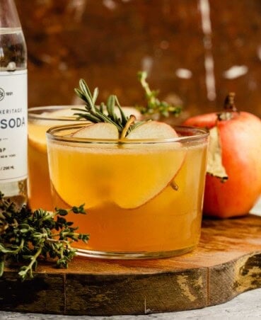 Short cocktail glass filled with an apple cider cocktail and garnished with an apple slice ad rosemary sprig. Cocktail set on a wood platter with an apple, bottle of club soda and more rosemary set around it.