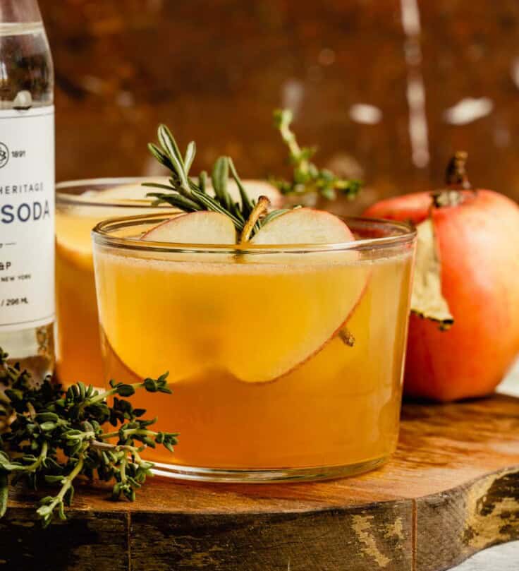 Short cocktail glass filled with an apple cider cocktail and garnished with an apple slice ad rosemary sprig. Cocktail set on a wood platter with an apple, bottle of club soda and more rosemary set around it.