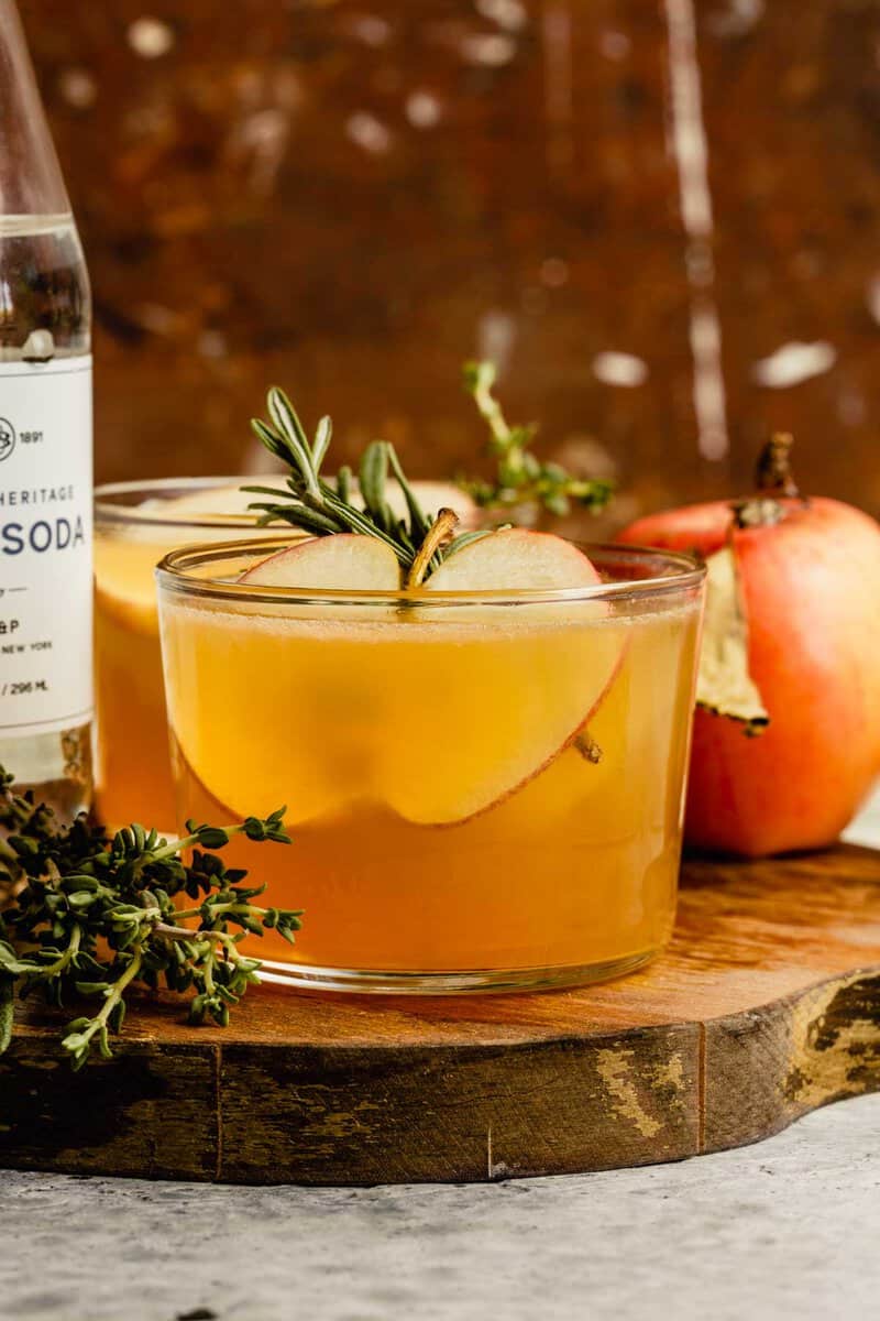 Short cocktail glass filled with an apple cider cocktail and garnished with an apple slice ad rosemary sprig. Cocktail set on a wood platter with an apple, bottle of club soda and more rosemary set around it.