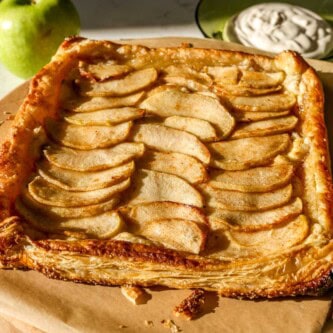 Golden brown baked apple puff pastry tart on parchment paper with apple slices neatly shingled and the edges folden up over the apples. Green apple and sweetened cream sauce set in the background.