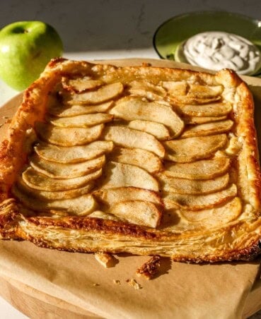 Golden brown baked apple puff pastry tart on parchment paper with apple slices neatly shingled and the edges folden up over the apples. Green apple and sweetened cream sauce set in the background.