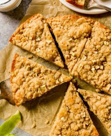 Apple tea cake with streusel on top set on a piece of brown parchment paper, cut into slices, with apple peels, milk and a plate set off the side.