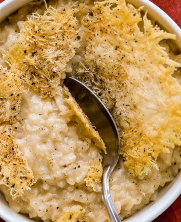 Cheese crisp with a spoon shattering through it into a creamy risotto in a shallow white bowl.