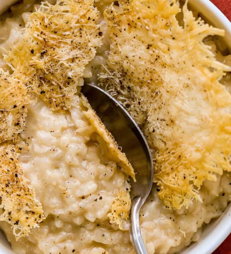 Cheese crisp with a spoon shattering through it into a creamy risotto in a shallow white bowl.