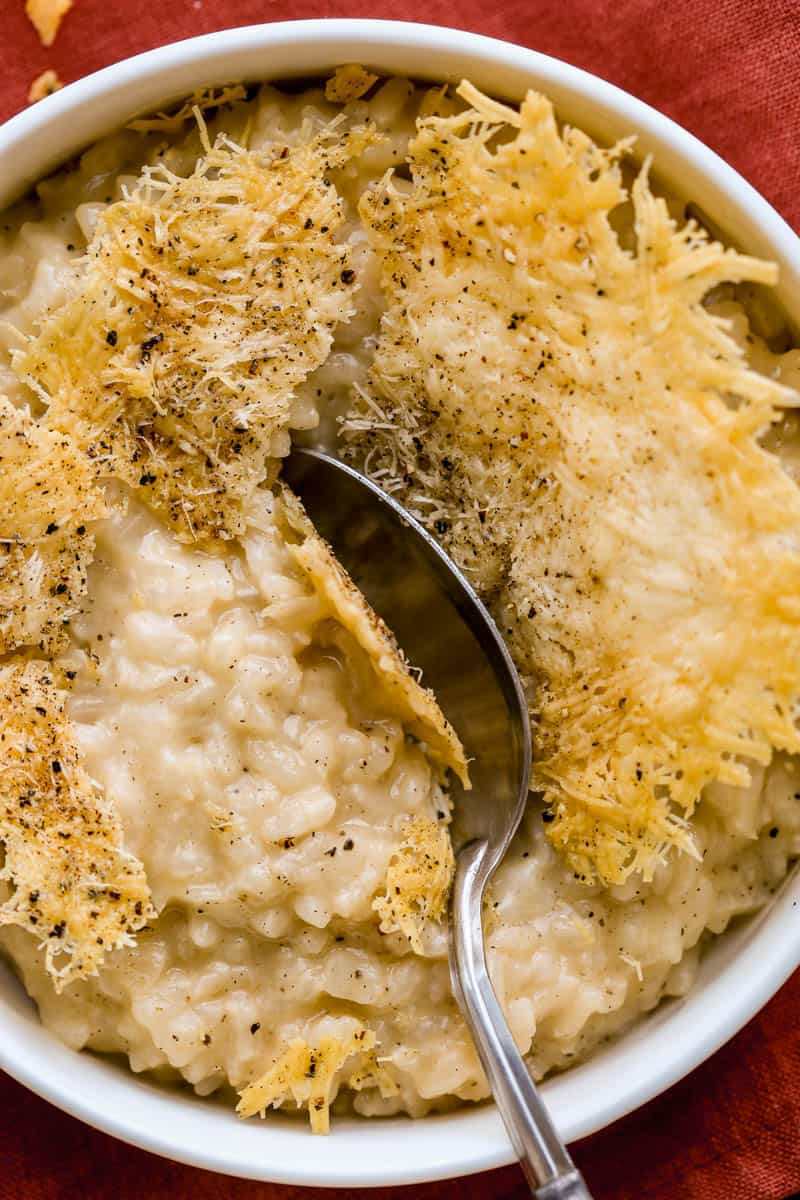 Cheese crisp with a spoon shattering through it into a creamy risotto in a shallow white bowl.