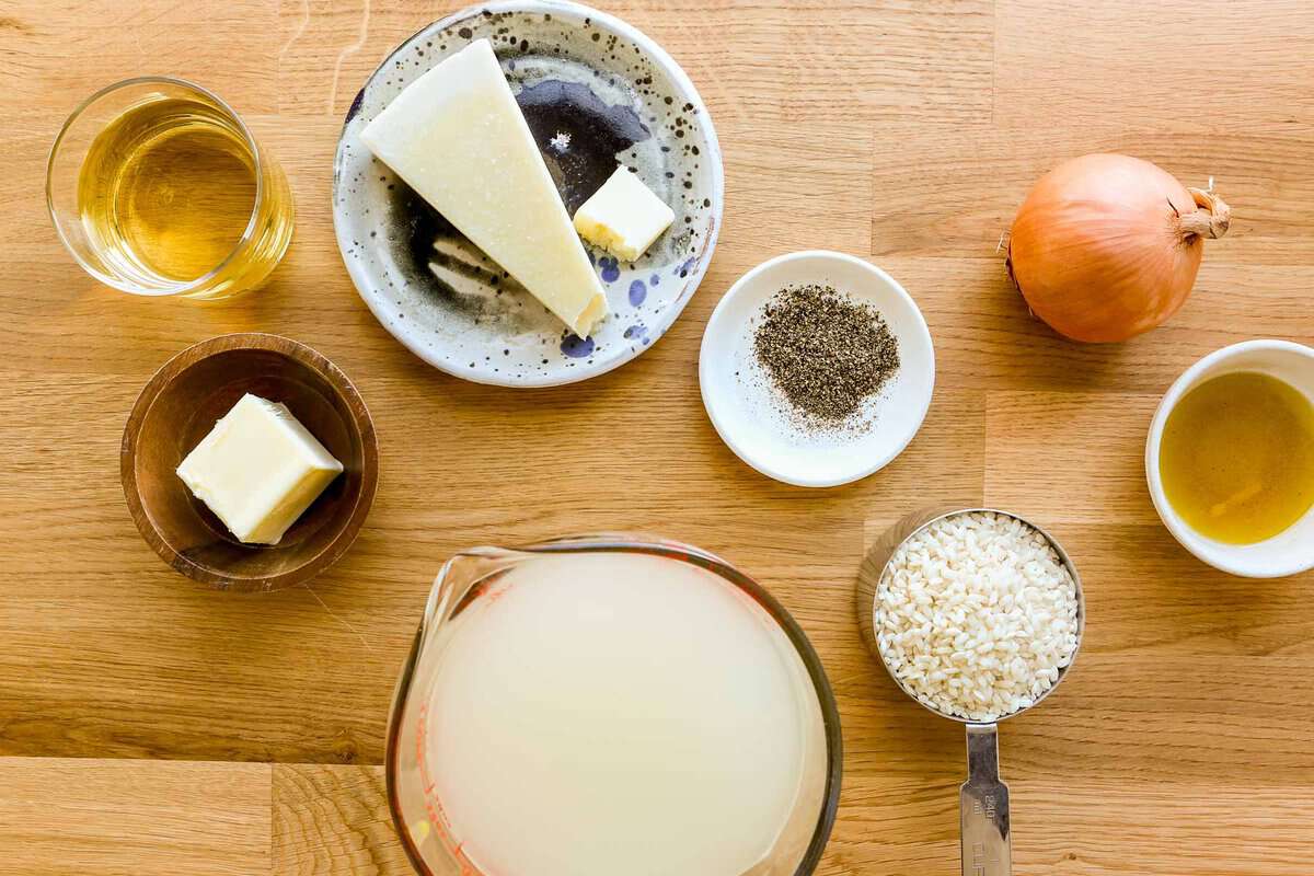 white wine, butter, pecorino cheese, broth, black pepper, arborio rice, onion and oil set out on a wood counter.