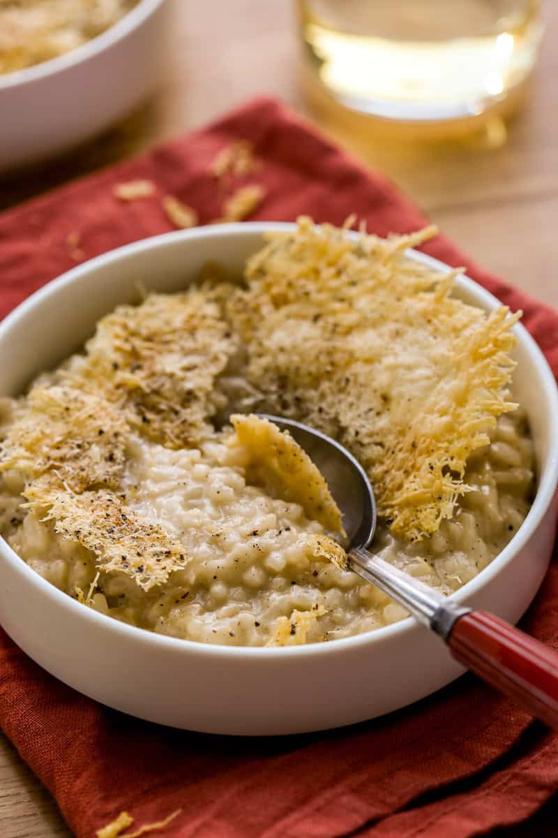 Cheese crisp with a spoon shattering through it into a creamy risotto in a shallow white bowl with a glass of wine set in the background.