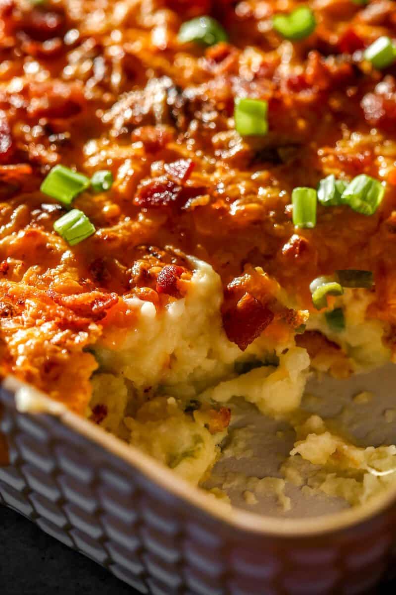 Baked mashed potatoes in a baking dish topped with melted and golden brown cheese, chopped scallions and bacon bits.