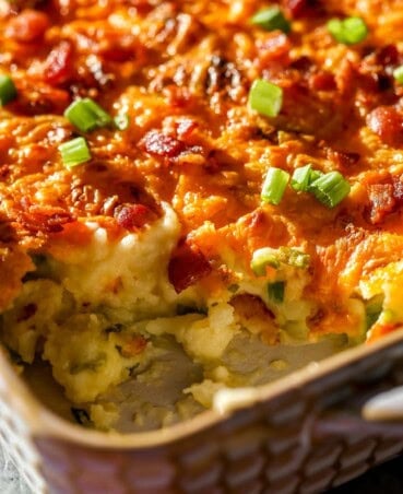 Baked mashed potatoes in a baking dish topped with melted and golden brown cheese, chopped scallions and bacon bits.