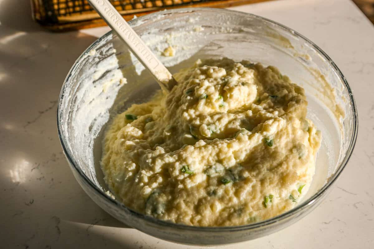 Mashed potato mixture with chopped scallions in a glass mixing bowl with a white spatula set in it.