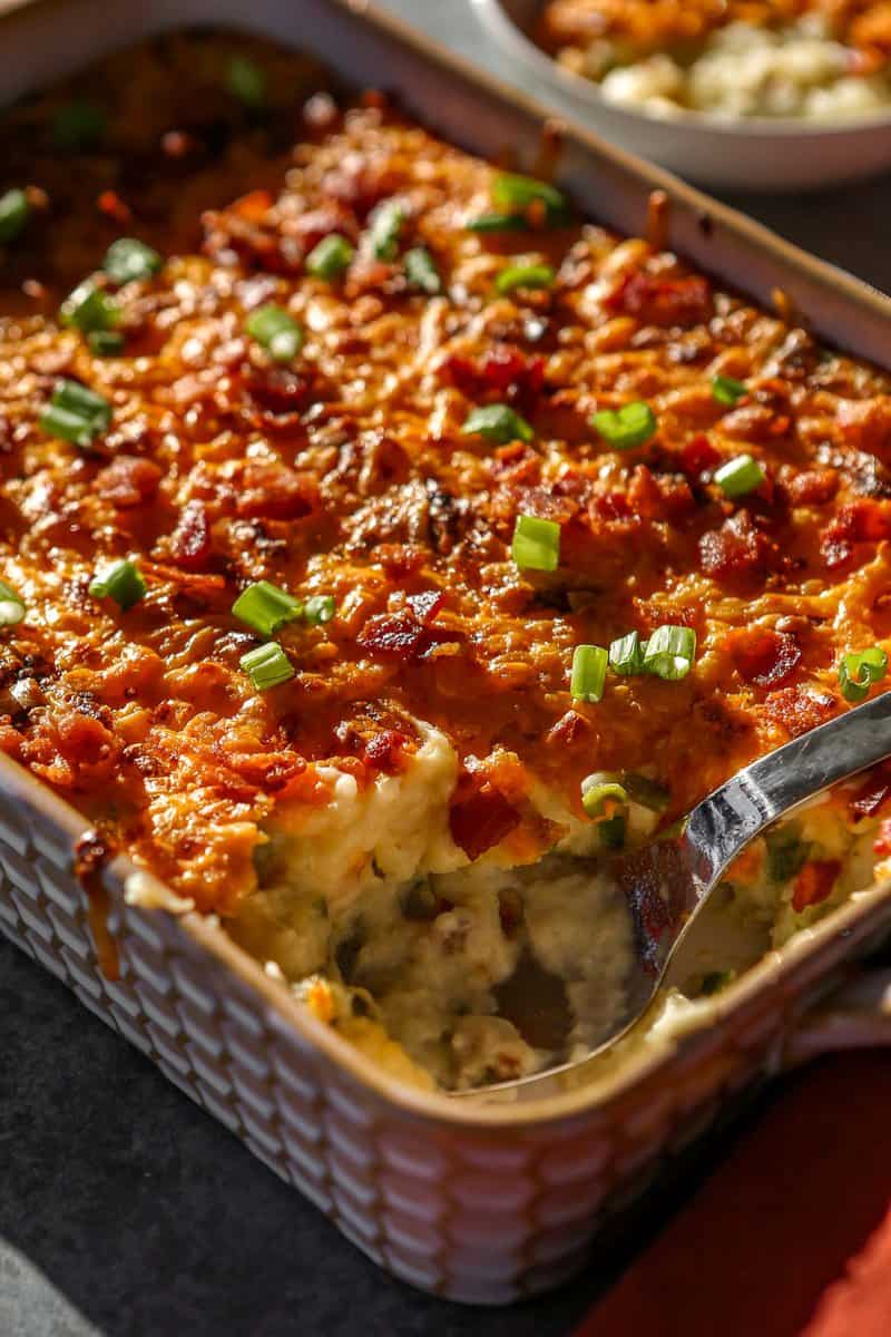 Baked mashed potatoes in a baking dish topped with melted and golden brown cheese, chopped scallions and bacon bits.