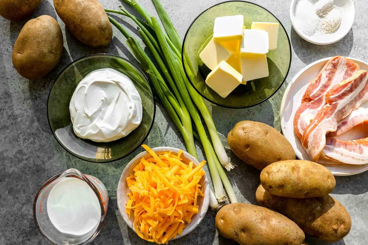 Russet potatoes, sour cream, milk, shredded Cheddar cheese, scallions, butter, uncooked bacon slices and garlic powder, onion powder and salt measured out and set on a counter.