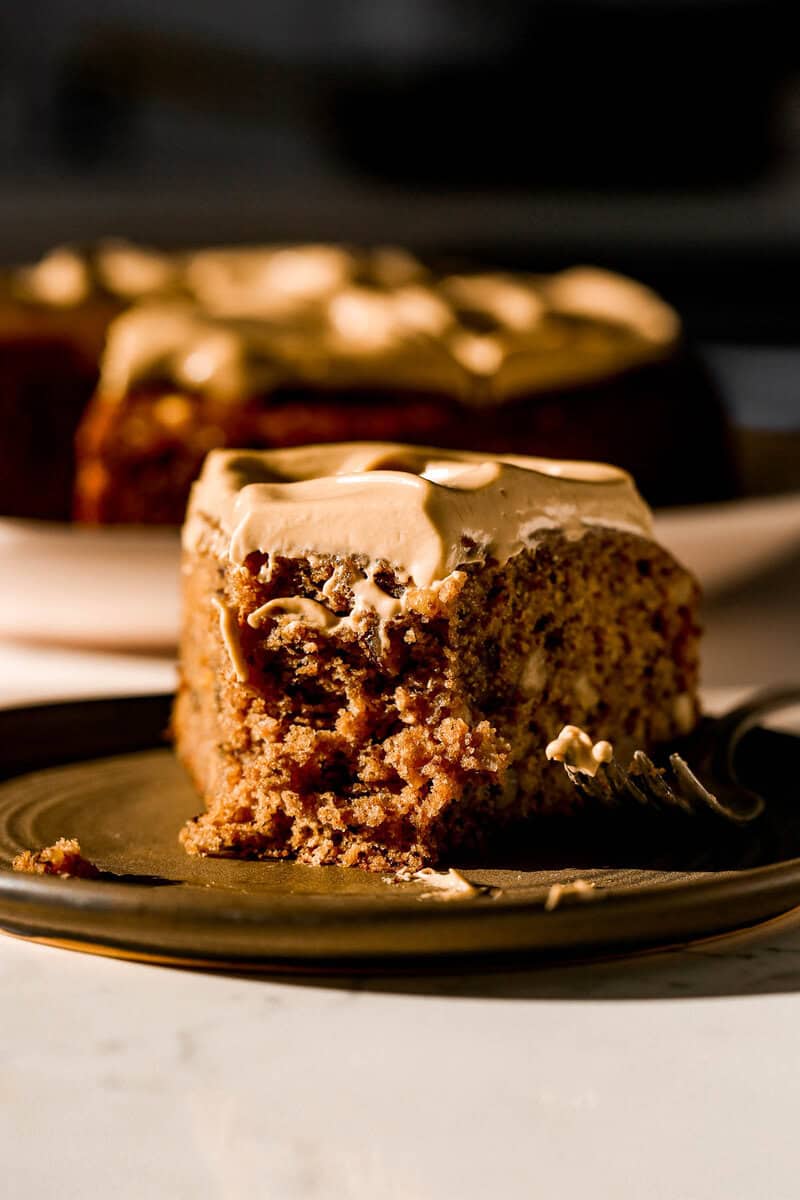 A close-up of a slice of date and walnut cake with a moist, textured crumb, topped with thick, smooth cream cheese frosting. The cake sits on a dark plate, with a fork placed beside it. Sunlight highlights the cake's golden-brown color and the creamy frosting, with the remaining cake blurred in the background.