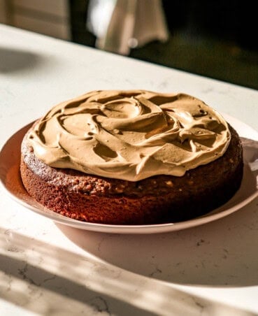 A full date and walnut cake with a rich, golden-brown crumb, topped with thick, swirled cream cheese frosting. The cake sits on a white plate, placed on a marble countertop with soft sunlight streaming across the scene, creating shadows and highlights that emphasize the smooth texture of the frosting.