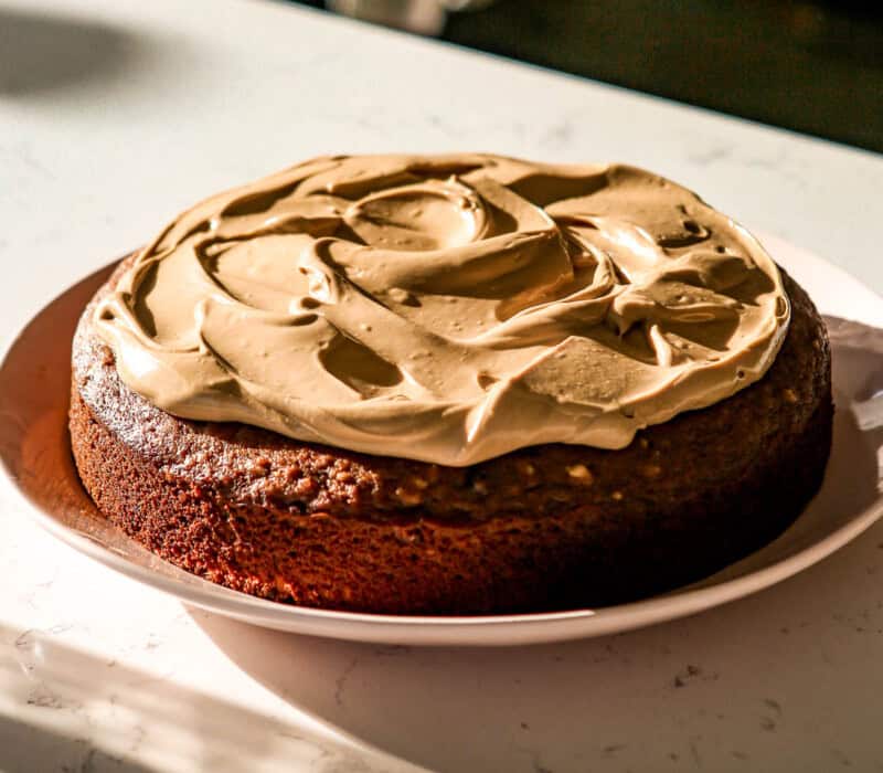 A full date and walnut cake with a rich, golden-brown crumb, topped with thick, swirled cream cheese frosting. The cake sits on a white plate, placed on a marble countertop with soft sunlight streaming across the scene, creating shadows and highlights that emphasize the smooth texture of the frosting.