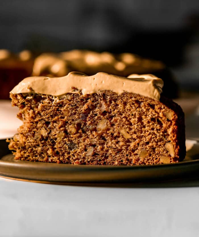 A close-up side angle of a slice of date and walnut cake with a moist, textured crumb, topped with thick, smooth cream cheese frosting. The cake sits on a dark plate. Sunlight highlights the cake's golden-brown color and the creamy frosting, with the remaining cake blurred in the background.