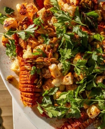 Hasselback-style cut roasted honeynut squash on a large white platter topped with cubes of broiled cheese, torn bread, pepitas and parsley.