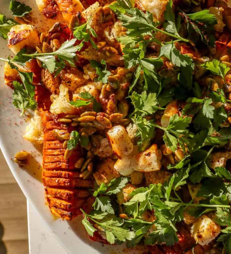 Hasselback-style cut roasted honeynut squash on a large white platter topped with cubes of broiled cheese, torn bread, pepitas and parsley.