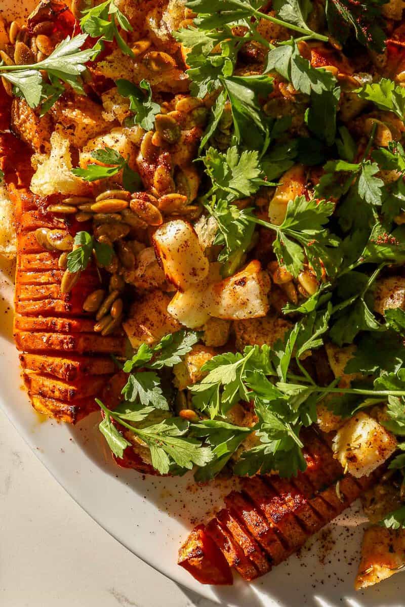 Hasselback-style cut roasted honeynut squash on a large white platter topped with cubes of broiled cheese, torn bread, pepitas and parsley.