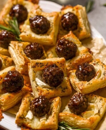 Meatball puff pastry bites made with glazed meatballs on top of baked puff pastry squares and melty brie cheese. Stacked on a large white platter with rosemary set around.