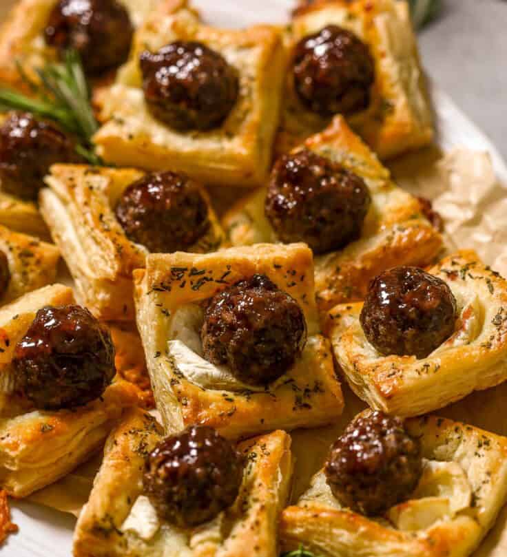 Meatball puff pastry bites made with glazed meatballs on top of baked puff pastry squares and melty brie cheese. Stacked on a large white platter with rosemary set around.