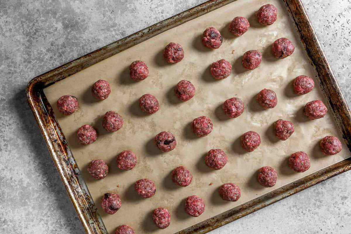 Uncooked beef meatballs arranged in even rows on a parchment lined baking sheet.