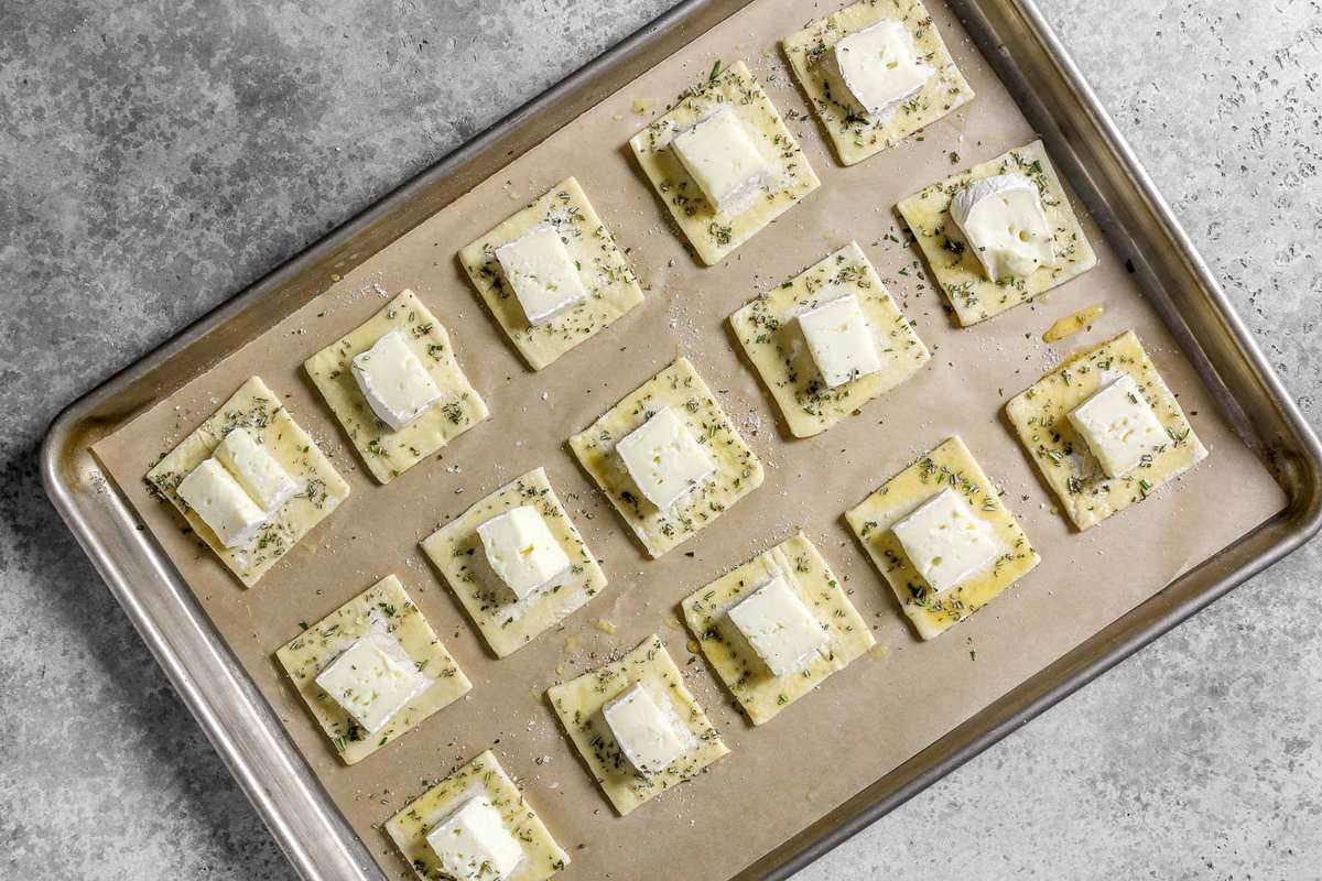 Unbaked puff pastry squares topped with a cube of brie cheese and minced fresh rosemary arranged in even rows on a parchment lined baking sheet.