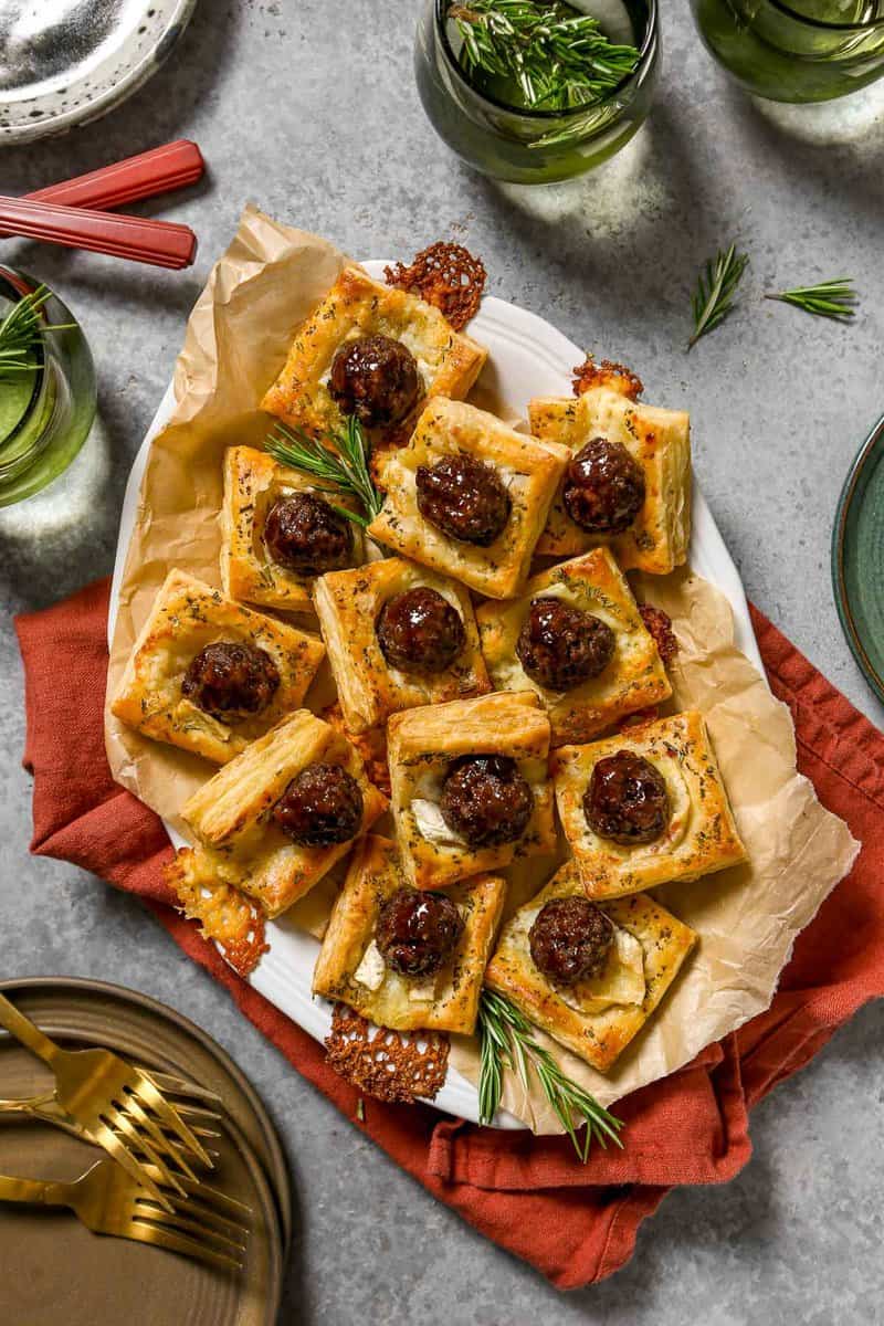 Meatball appetizer bites made with glazed meatballs on top of baked puff pastry squares and melty brie cheese. Stacked on a large white platter with rosemary set around.