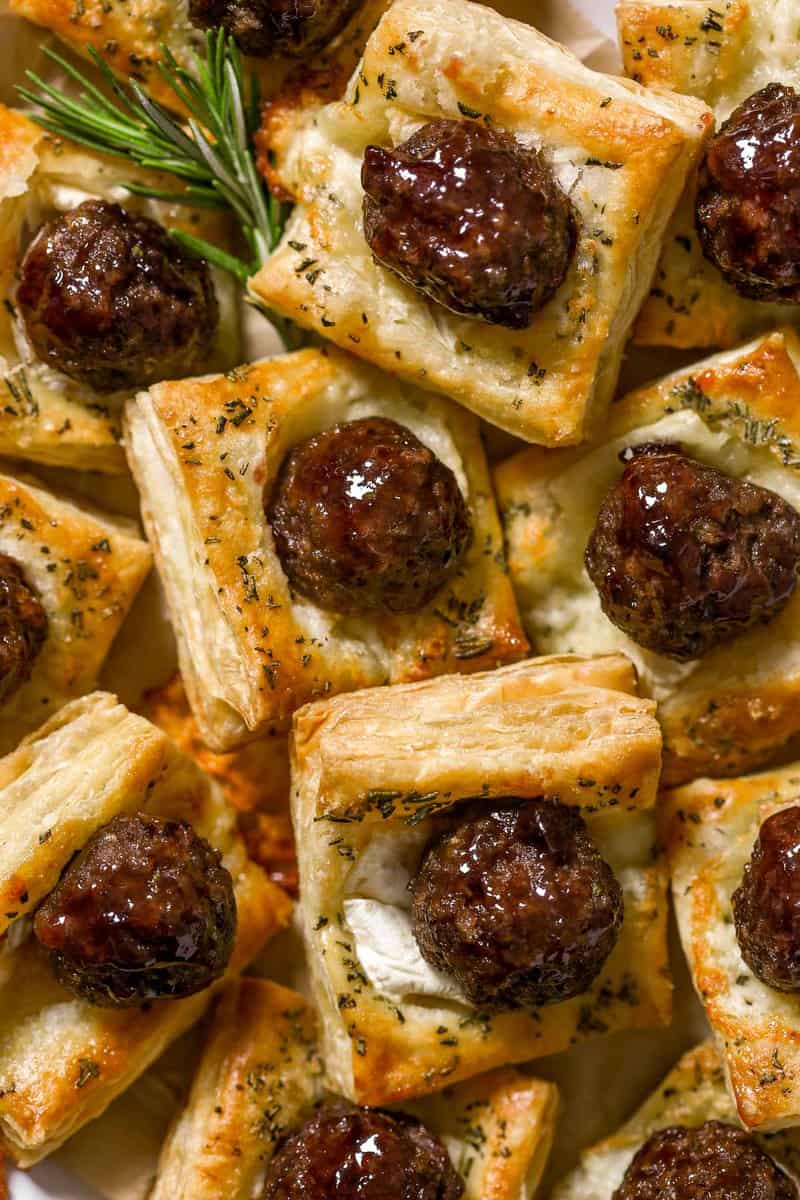 Meatball puff pastry bites made with glazed meatballs on top of baked puff pastry squares and melty brie cheese. Stacked on a large white platter with rosemary set around.