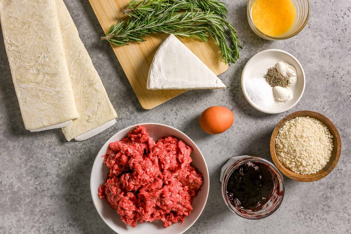 Two folded sheets of puff pastry, ground beef, fresh rosemary, wedge of brie cheese, egg, jam, breadcrumbs, spices and beaten egg in a small glass bowl all set out on a counter.