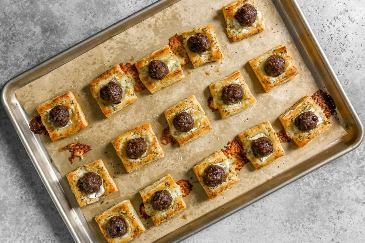 Parchment lined baking sheet with baked meatball appetizer bites, made with glazed meatballs on top of baked puff pastry squares and melty brie cheese, arranged in even rows.