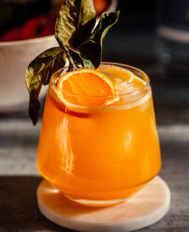 Vibrant orange cocktail in a rocks glass garnished with a slice of clementine. Cocktail set on a white coaster.